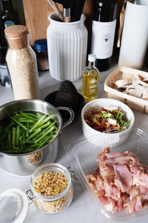 Kitchen countertop covered in different meal prep items, including green beans, chicken thighs, grains, and mushrooms, as well as a prepped ramekin of food.