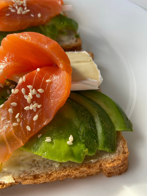 Two slices of toast topped with avocado, brie, salmon, and sesame seeds.
