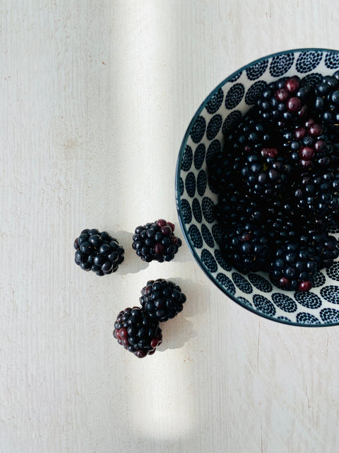 Overhead view of a bowl of blackberries on a tabletop, with 4 blackberries strewn across the table next to the bowl.