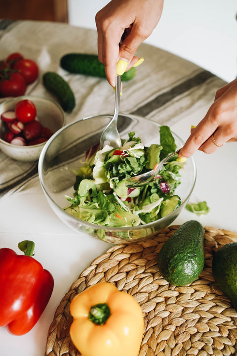 Keto meal prep, with woman tossing a salad in a glass bowl on a countertop covered in fresh vegetables and with a bowl of sliced radishes in the background.