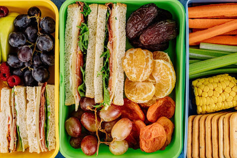 Prepped meals in reusable containers, with sandwiches, dates, grapes, corn, celery, crackers, clementines, and carrots.