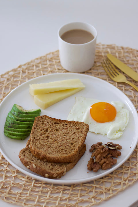 Breakfast placement with sliced avocado, 2 slices of cheese, 2 slices of bread, an over easy egg, and some nuts, next to a cup of coffee.