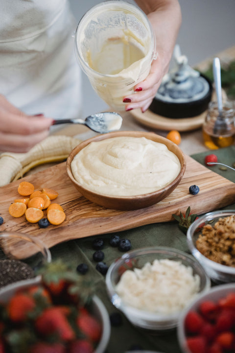 Person prepping a smoothie bowl, pouring the fruit puree into a bowl on a cutting board, surrounded by bowls of toppings, including strawberries, coconut flakes, nuts, and raspberries.