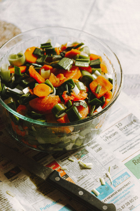 Vegetarian meal prep in a bowl, with carrots, legumes, and green onions. The bowl sits atop a countertop with a newspaper and a knife.