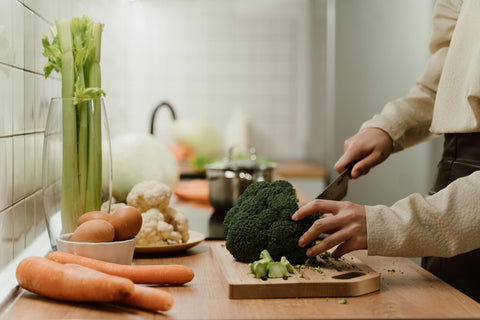 Person chopping broccoli on a cutting board surrounded with other produce, including carrots, eggs, cauliflower, and celery.